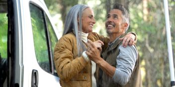 Moden kvinde og mand danser smilende i regnen uden for deres autocamper. Bag dem kan man se træer. 