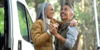 Moden kvinde og mand danser smilende i regnen uden for deres autocamper. Bag dem kan man se træer. 