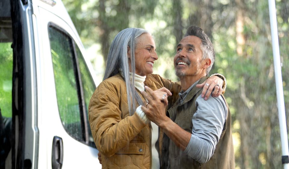 Moden kvinde og mand danser smilende i regnen uden for deres autocamper. Bag dem kan man se træer. 