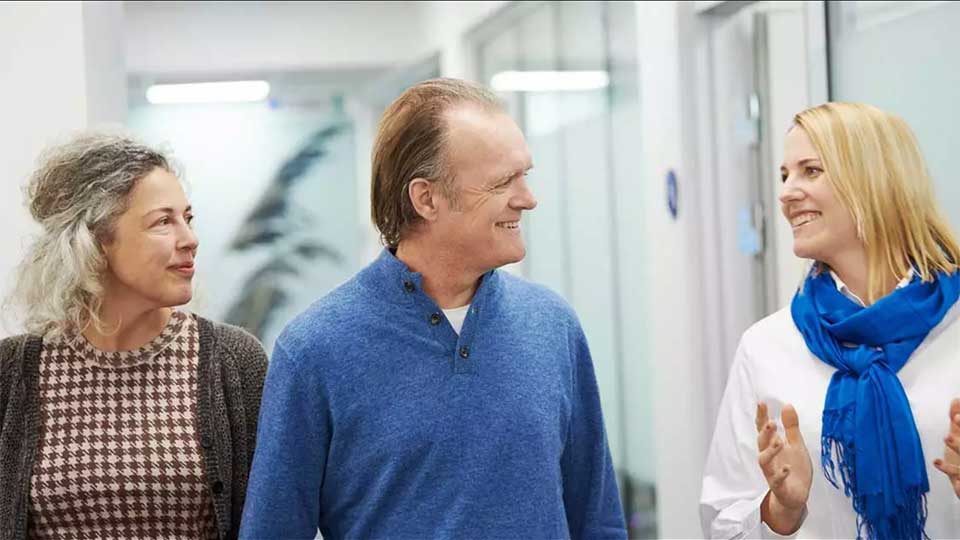 Tre personer går i en moderne høringklinik med lyse vægge og glasdøre; en audiolog med blå tørklæde kommunikerer med en mand i blå trøje og en kvinde ved siden af.