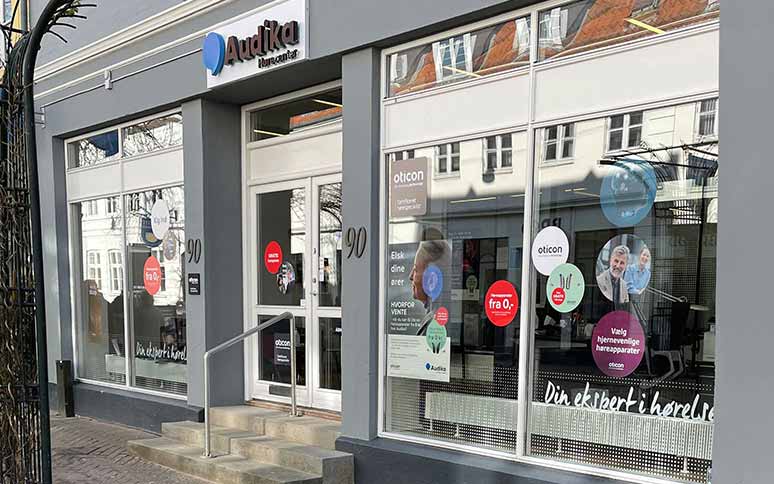 Audika-høreklinik med glasfacade viser reklamer for Oticon-høreapparater, herunder teksten ’Elsk dine ører’ og ’fra 0,-’. Bygningen har en hoveddør og adressemarkering ’90’.