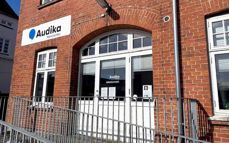 Audika Hørecenter-bygning i røde mursten med en hvid indgangsdør. Skiltet på facaden viser 'Audika Hørecenter'. Indgangen er tilgængelig via en rampe foran døren.