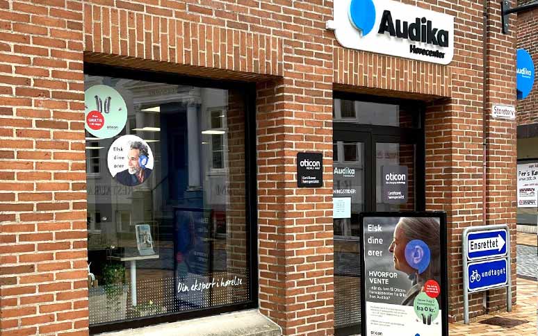 Audika hørecenter med murstensfacade viser reklamer for Oticon-høreapparater i vinduerne med teksten 'Elsk dine ører' og 'Din ekspert i hørelse.' Udendørs skilte informerer om høretests og produkter.