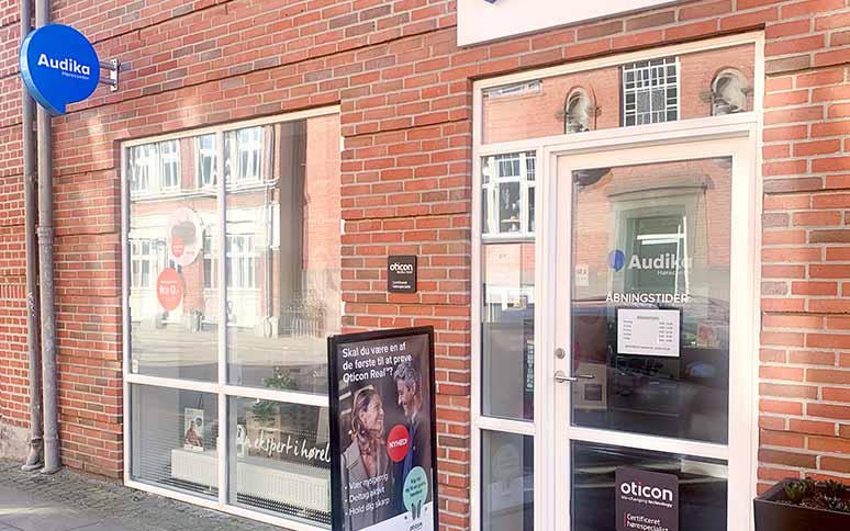 Audikas høreklinik med murstensfacade, Oticon-skilt og vinduer med tekst som 'Få det bedste ud af din hørelse.' En plakat reklamerer for Oticon Real høreapparater. Åbningstider ses på døren.