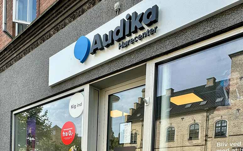 Audika Hørecenter-facade viser logo med blå ikon og tekst, vindue med klistermærker: 'Kig ind,' 'Høreapparater fra 0 kr.*', beliggende i byområde foran murstensbygning.