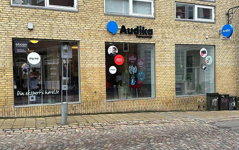 Audika hørecenterfacade med gule mursten, store vinduer og skilte med reklamer for gratis høretest, Oticon-høreapparater og mottoet 'Din ekspert i hørelse.' Beliggende ved brostensbelagt fortov.