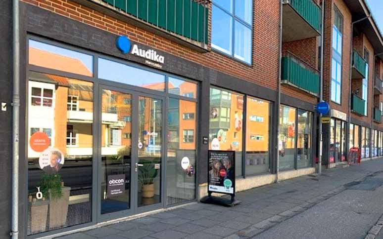 Audikas høreklinik med glasdøre og vinduer viser deres logo, reklametekster og Oticon-brandet. Klinikfacaden er placeret i en moderne bygningsgade omgivet af rød murstensarkitektur.