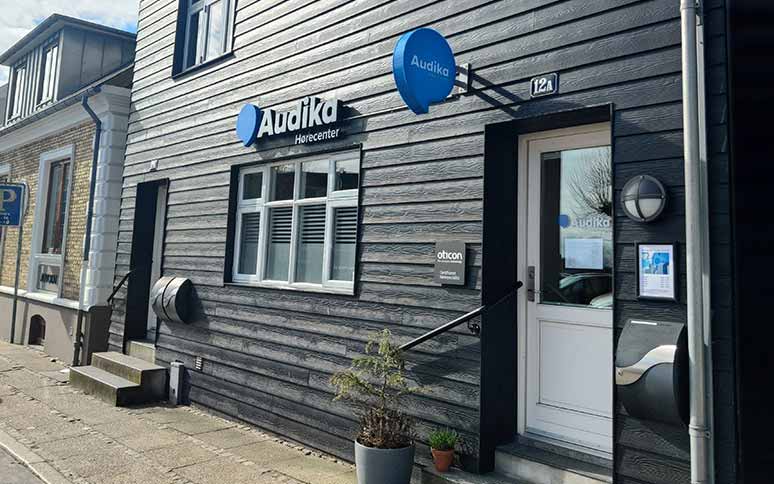 Audika Hørecenter-butik med sort træfacade og logo i blåt, placeret på en hyggelig gade. Skilt nævner Oticon. Indgang til høreklinik via hvid dør med trappe.