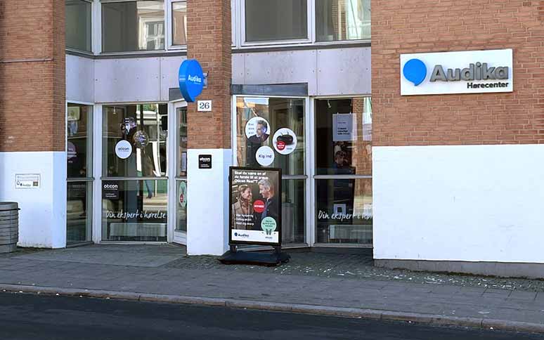 Audika Hørecenter-indgang med murstensbygning; skilte viser Oticon-høreapparater og tekst inklusive ’Din ekspert i hørelse’ og ’Kom ind.’ Reklameskilt foran fokuserer på høretest og audiologi.