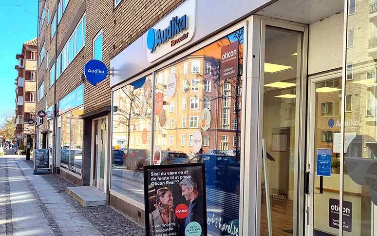 Audika-hørecenter med glasfacade viser reklamer for høreapparater fra Oticon, herunder 'Oticon Real' med teksten: 'Skal du være en af de første til at prøve Oticon Real?'. En gågade med bygninger ses i baggrunden.