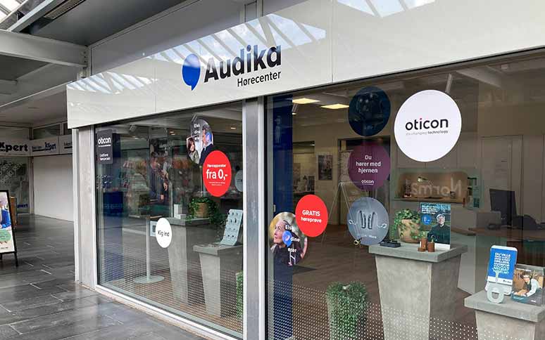 Audika Hørecenter, facade med glasvinduer, reklamerer for høreapparater fra Oticon. Tekst om gratis høreprøve og tilbud 'fra 0 kr' ses tydeligt; indvendigt vises høreklinikens reception og displays.