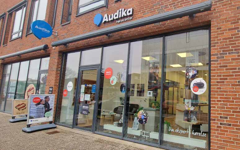 Audika Hørecenter butikfacade med glasvinduer, murstensbygning og skilte for høreapparater (Oticon nævnt). Tekst: 'Din ekspert i hørelse' og reklamer for høretests. Indvendigt ses møbler og brochurer.