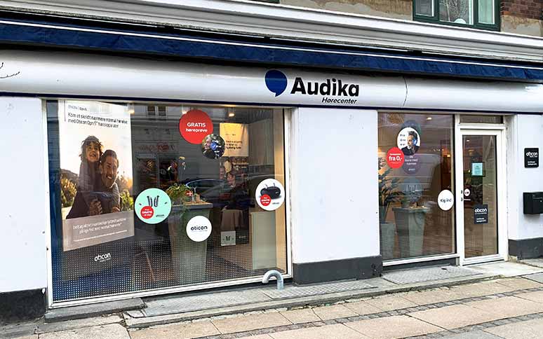 Audika Hørecenter-facade med glasvinduer, reklamer for Oticon-høreapparater, tekst som ’GRATIS høreprøve’ og ’fra 0 kr. med offentlig tilskud’, omgivet af bymiljø og flisebelagt fortov.