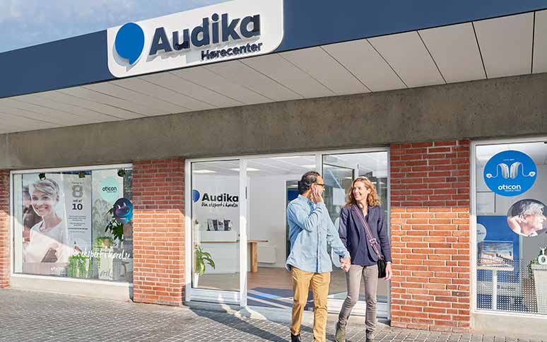 Et par går hånd i hånd foran Audikas hørecenter med en murstensfacade. Skilte viser Oticon-brandet, og vinduer reklamerer for høreapparater og høretest inde i klinikken.