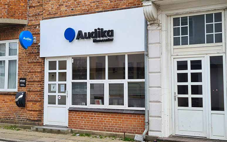 Facaden af en Audika-høreklinik med et hvidt skilt, hvorpå der står 'Audika Hørecenter' i sort tekst. Bygningen er af røde mursten med hvide vinduer.