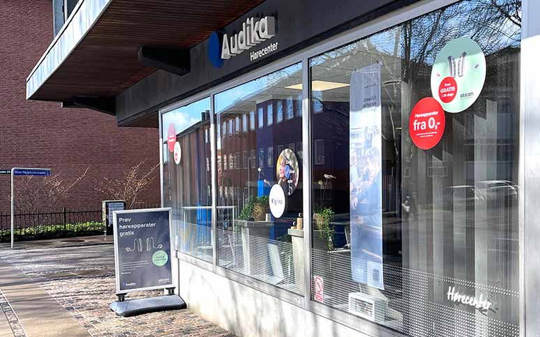 Audika Hørecenter-butikken har store glasvinduer med reklamer for høreapparater, inkl. Oticon. Et gadeskilt tilbyder gratis prøve af høreapparater. Butikken ligger ved en flisebelagt gade i solskin.
