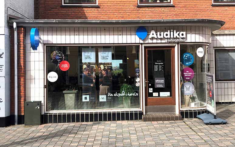 Audika Hørecenter-facade med glasvinduer viser skiltning og produkter. Tekst på vinduer inkluderer ’Din ekspert i hørelse’ og reklame for Oticon-høreapparater. Muret bygning i bymiljø.