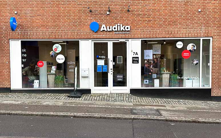 Audika Hørecenter facade med røde mursten, store vinduer og tekst som 'Kig ind,' 'Oticon,' og 'Din lokale høreekspert.' Indenfor vises brochurer og høretest-relateret udstyr.