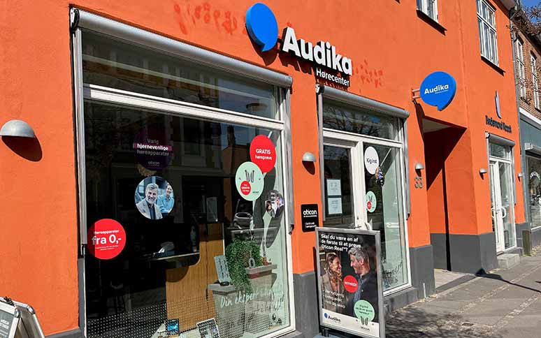 Audika Hørecenter-butik med orangerod facade og store vinduer fyldt med reklamer. Tekst inkluderer: ’GRATIS høreprøve,’ ’fra 0,-,’ og ’Oticon.’ Skilte promoverer høreapparater og høretests.