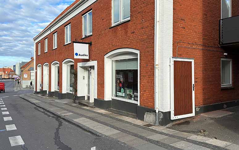 Rødt murstensbygning med skiltet ’Audika’ over indgangen. Klinikken tilbyder høretests og rådgivning om høreapparater. Placering ved fortovet i en bymæssig gade med parkerede biler.