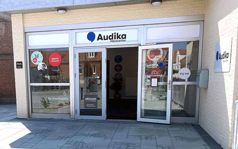 Audika Hørecenter-indgang med glasdøre, reklamer for ’GRATIS høreprøve’ og Oticon-høreapparater. Bygningen er omgivet af kultiverede fliser og mursten, tydeligt en professionel høreklinik.