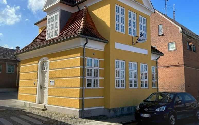 En gul bygning med hvide vinduer og skiltet ’Audika’ på facaden huser en høreklinik. En sort bil holder parkeret foran på en brolagt gade under blå himmel.