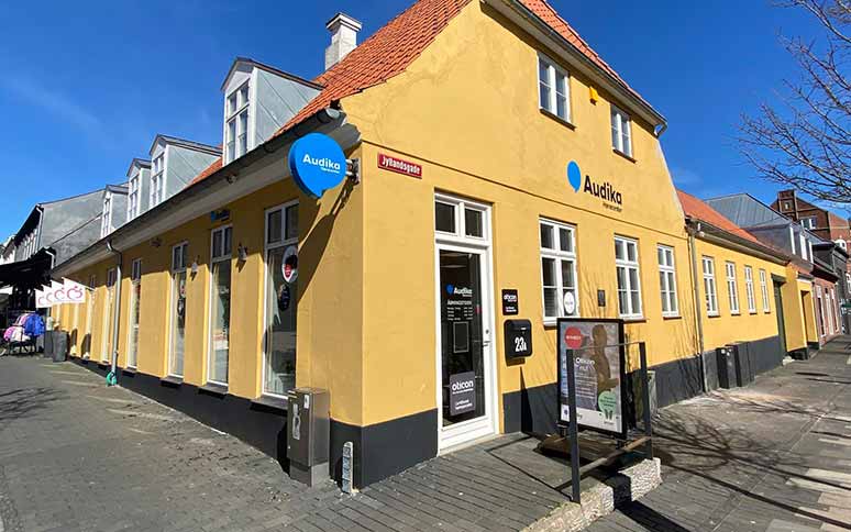 Audika-høreklinik i en gul bygning med rødt tag på Jyllandsgade 23A. Skilte viser ’Audika,’ ’Oticon’ og ’Høretest.’ Solrig dag med træer og gadebutikker omkring.