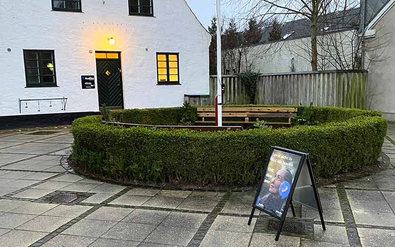 En hvid bygning med grønne døre og vinduer. Foran ses en hæk omringende en bænk og et skilt. Skiltet viser tekst: ’Hør øjeblikkene, du vil huske resten af livet,’ og et billede af en mand med høreapparat. Audika-logo og Oticon-brand er synlige.