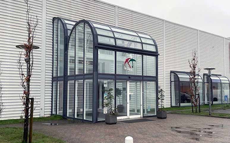 Bygningens glasskydedør i buet indgang er dekoreret med KS Gruppen-logoet; omgivet af grå facadepaneler, unge træer og vådt fortovsbelægning under en overskyet himmel.