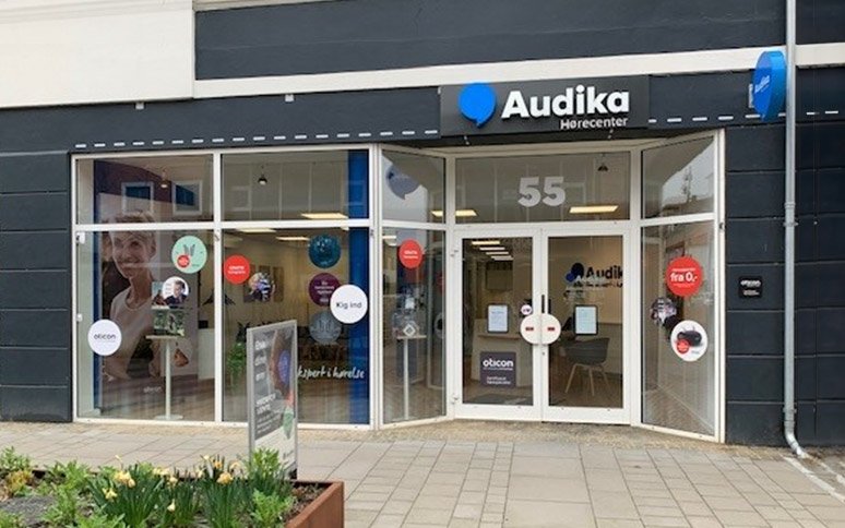 Audika Hørecenter med facade ud mod en fortovsplads. Skiltning viser ’Audika’ og adresse ’55.’ Vinduesdekorationer inkluderer Oticon-logo, tilbud fra 0 kr., og tekst som ’Kig ind’ og ’Eksperti hverdag.’