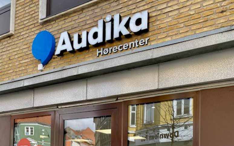 Audika Hørecenter-logo pryder facade af gul murstensbygning; hovedindgangen viser glasdøre reflekterende bymiljø. Tilbyder høreapparater, høretjek og audiologisk rådgivning for forbedret hørelse.