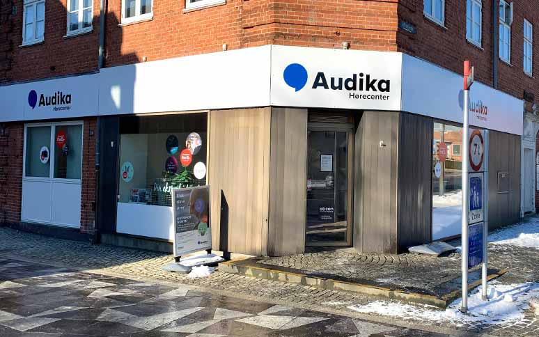 Audika Hørecenter-butik med logo på facade ligger på et hjørne af en murstensbygning. En reklameskilt udenfor promoverer Oticon-høreapparater. Omkring er sne og brostensbelægning.