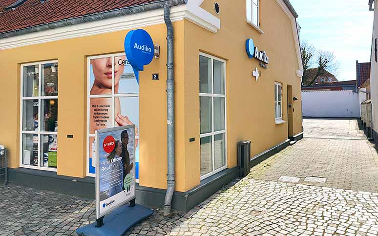 Audika-høreklinik i en gul bygning med blå skiltning. En sandwichplakat udenfor reklamerer for ’Nyhed: Prøv Oticon Real nu!’ og fremhæver løsninger til bedre høreoplevelser. Flisebelagt gade omkring.