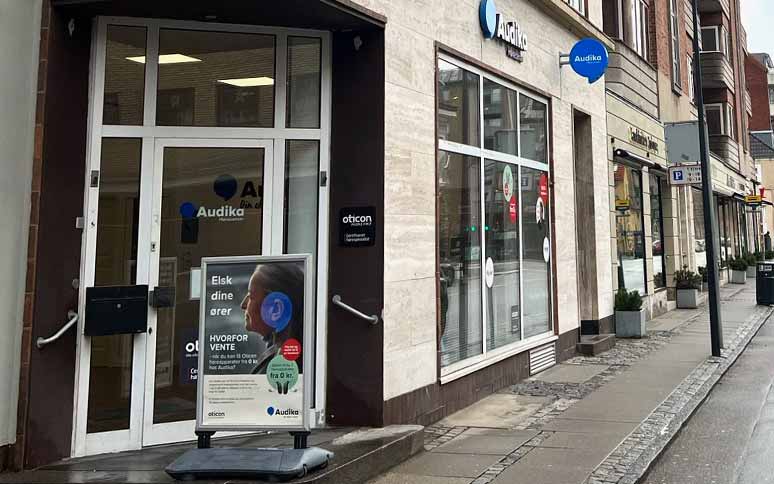 Audika-høreklinik med facadevinduer og dør, skilt reklamerer for Oticon-høreapparater med teksten: ’Elsk dine ører. HVORFOR VENTE. Få en tid til gratis høretest i dag.’ gadeomgivelse.