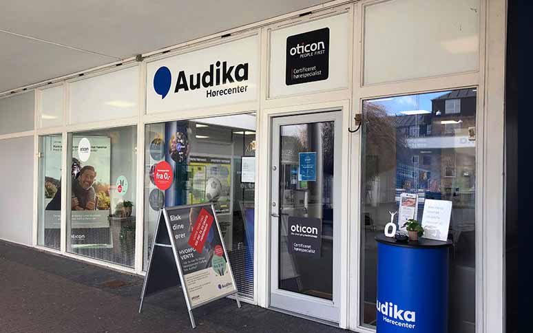 Audika Hørecenter-butik med glasfacade og synlige reklameskilte for Oticon høreapparater. En sandwichstander foran indgangen promoverer høretest. Indgangen leder ind i en moderne høreklinik i bymiljøet.