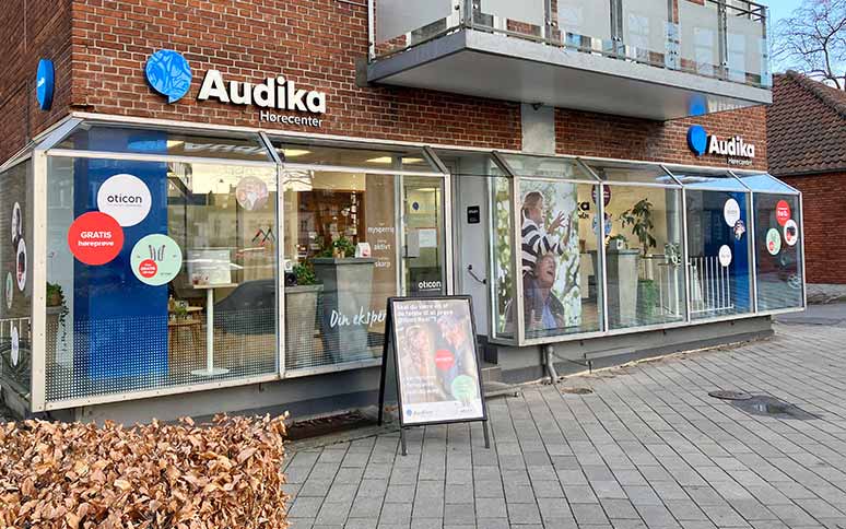 Audika Hørecenter-butik med glasfacade og murstensvæg viser deres logo og plakater for høreapparater fra Oticon. Stående skilt reklamerer for gratis høreprøvninger. Beliggende på en flisebelagt gade.
