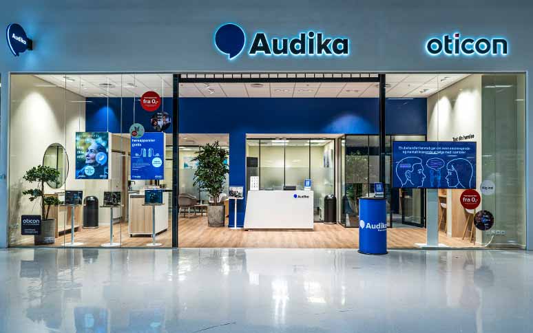 Audikas moderne høreklinik med glasfacade, blå indretning og godt oplyst receptionsområde. Oticon-logo vises; fokus på høreapparater, høresundhed og professionelle audiologiske tjenester. Infografik og reklamer om hørepleje.