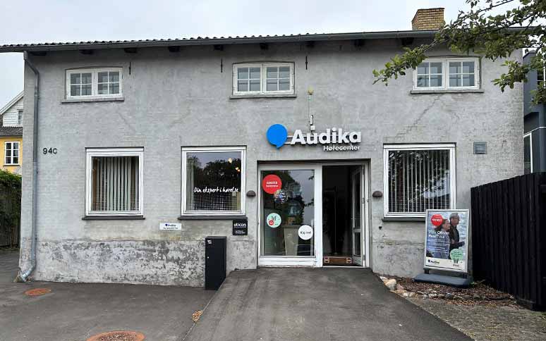 Audika Hørecenter-bygning med et skilt på facaden, der inkluderer ’Audika’-logoet. Døren fører til en høreklinik; Oticon-logoet ses ved indgangen. Plakat reklamerer for høreapparater.