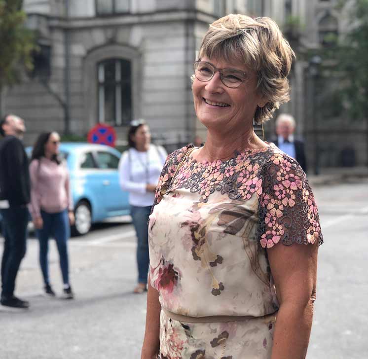 En kvinde iført en blomsterdesignet kjole står udendørs. Hun er omgivet af mennesker og en lyseblå bil i en urban kontekst med historiske bygninger i baggrunden. Ingen tekst synlig.