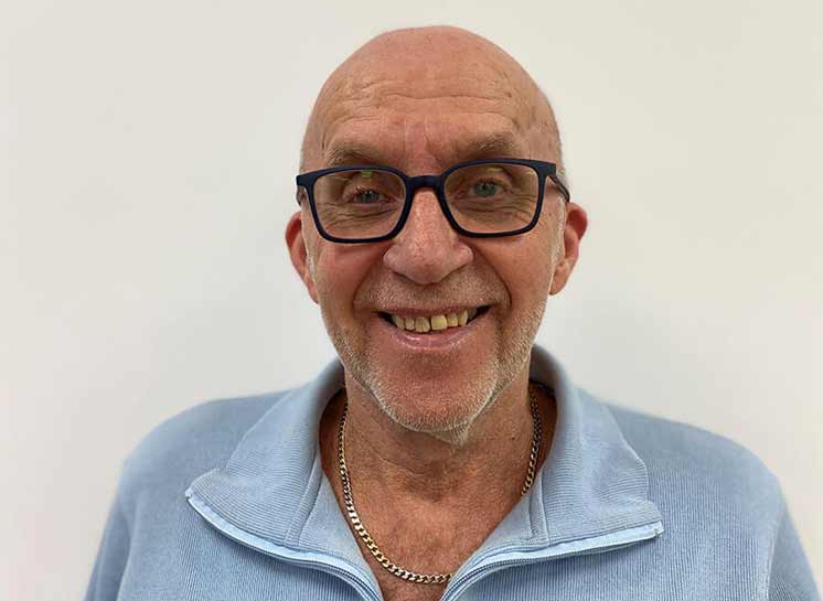 En ældre mand iført en lyseblå trøje med lynlås og en guldkæde om halsen, set mod en lys, neutral baggrund. Hans ansigt er sløret.