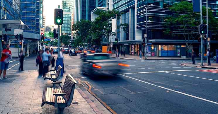 En central bykrydsning med travle fodgængere og biler i bevægelse. Forgrunden viser bænke langs fortovet, mens moderne bygninger omgiver området under tændte grønne lys. Ingen tekst identificeret.