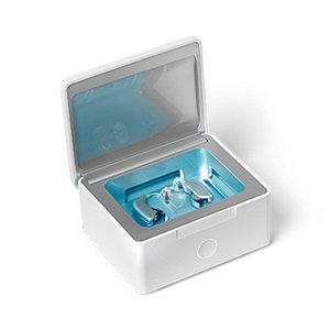 Hvid rengøringsboks med åbent låg, der indeholder to høreapparater under blåt UV-lys. Velegnet til rengøring og vedligeholdelse af høreapparater, typisk brugt i høreklinikker som Audika.