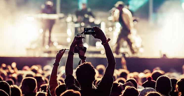Person i forgrund løfter smartphone for at optage en koncert, hvor musikere optræder på oplyst scene med røg og lys. Publikum fylder den mørkt oplyste forgrund. Ingen tekst synlig.