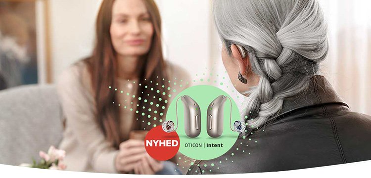 En ældre kvinde med gråt hår og fletning ses bagfra under en samtale med en yngre kvinde i en lys stue. Forgrunden viser to høreapparater fra Oticon, markeret med teksten ’NYHED OTICON | Intent.’ Reklamen refererer til høreklinikker og høreløsninger.