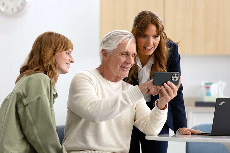 [GEN] An older man in a white sweater demonstrates a smartphone to two women in a modern office-like setting, with a Lenovo laptop and light wooden cabinetry in the background. No visible hearing aids or text.