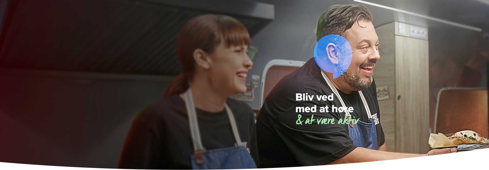 To personer iført forklæder arbejder i et køkkenmiljø; den ene holder en tallerken med mad. Tekst på billedet: ’Bliv ved med at høre & at være aktiv’.