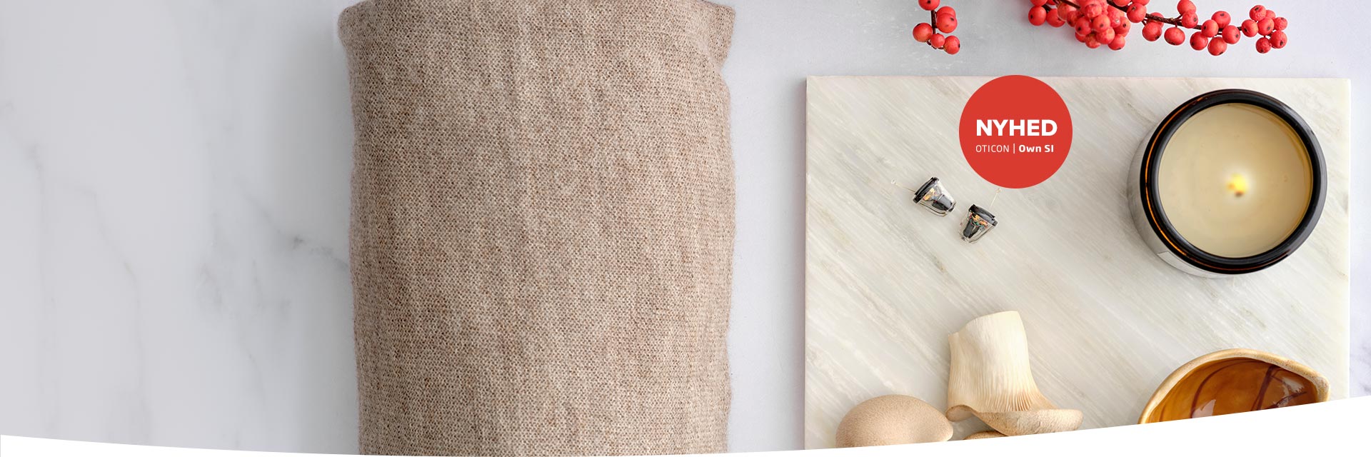 To høreapparater fra Oticon Own™ SI ligger på en lys overflade ved siden af en tændt lys, røde bær og naturlige dekorationer. Tekst: 'NYHED OTICON | Own SI'.