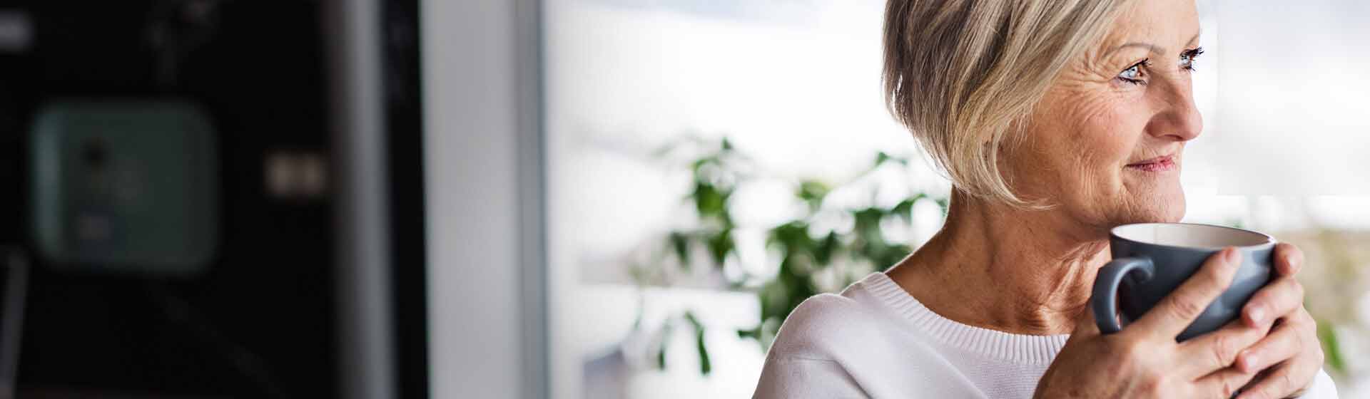 En ældre kvinde med kort, gråt hår holder en blå kop med begge hænder. Hun sidder indendørs i et lyst miljø med grønne planter i baggrunden.