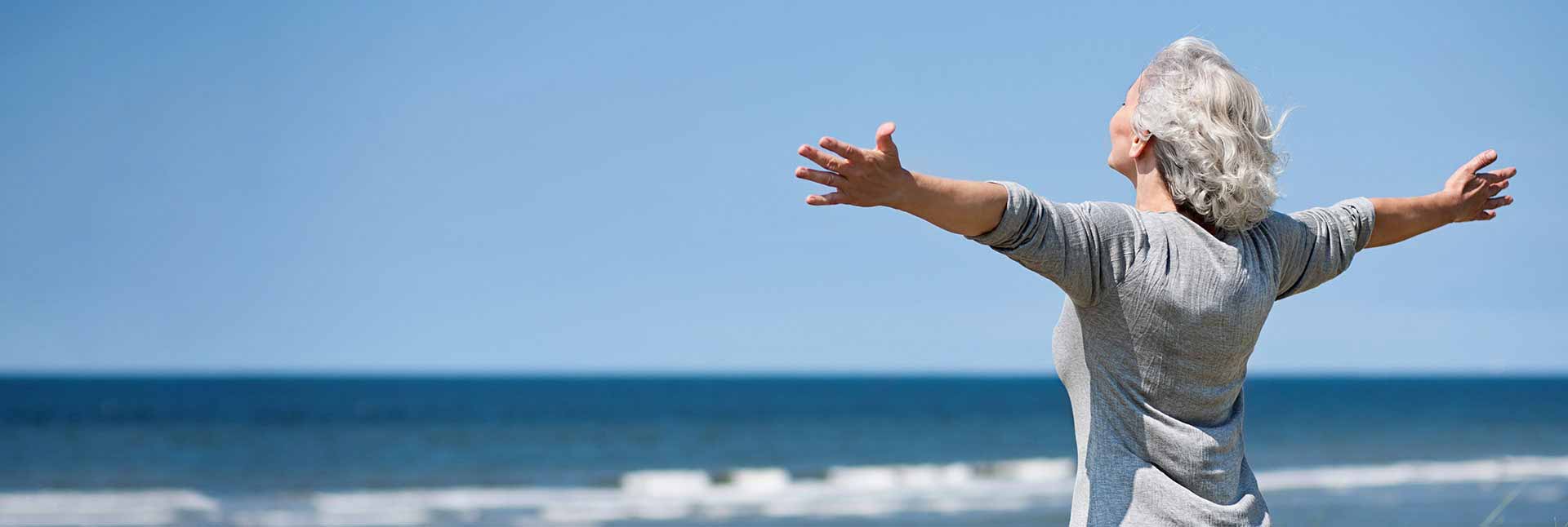 En ældre kvinde med gråt hår strækker armene ud mod havet, mens hun står på en strand under en blå himmel og nyder frihed og natur.