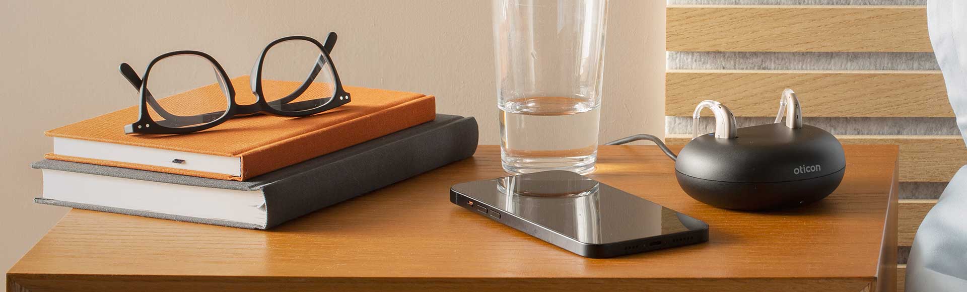 Tilbehør til Oticon høreapparater på natbord med oplader, briller, bøger, glas vand og smartphone